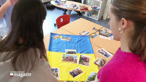 Zwei Schülerinnen betrachten eine ukrainische Flagge und daraufliegende Fotos. © Screenshot 