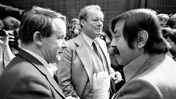 Siegfried Lenz im Gespräch mit Günter Grass, dahinter Willy Brandt (Bild von 1977) © Imago Foto: Sven Simon
