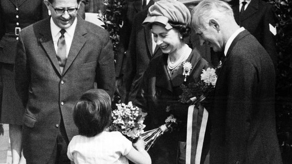 Ein kleines Mädchen überreicht Königin Elizabeth II. beim Verlassen des Bahnhofs Dammtor in Hamburg am 28. Mai 1965 einen Blumenstrauß. Links ein Dolmetscher, rechts Bürgermeister Paul Nevermann. © picture alliance / dpa 
