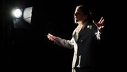 Eine junge Frau im grauen Anzug steht auf der Bühne im Licht eines einzelnen Scheinwerfers und schreit. © Landestheater Schleswig-Holstein Foto: Hernik Matzen