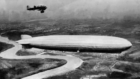 Ein Zeppelin fliegt 1928 über eine Landschaft begleitet von einem Doppeldeckerflugzeug. © dpa picture-alliance 