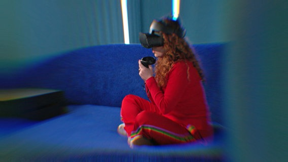 Eine Frau sitzt auf einem Sofa und trägt eine VR-Brille. © NDR/i&u Tv 