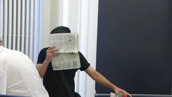 Ein Angeklagter verdeckt sein Gesicht mit einer Zeitung. © Screenshot 
