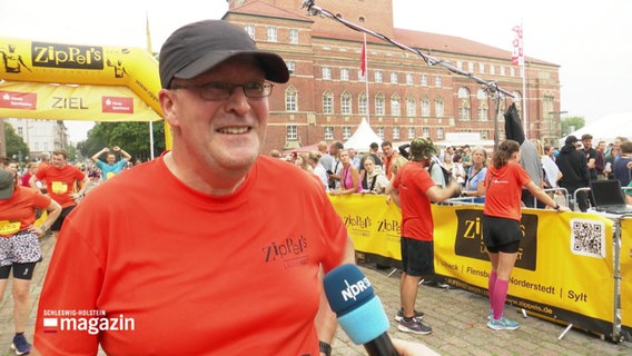 Ein lächelnder Mann mit rotem Shirt und Baseball-Kappe wird interviewt © Screenshot 