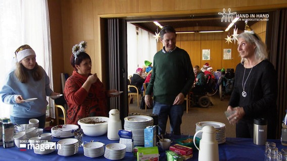 Drei Frauen und ein Mann stehen vor einem Büffet mit Tellern und Speisen. © Screenshot 