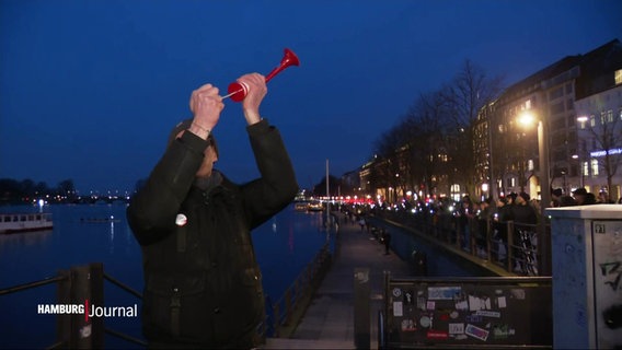 Ein Mann steht vor der Binnenalster und betätigt eine rote Tröte. Im Hintergrund sieht man eine Menschenkette mit Lichtern in den Händen. © Screenshot 