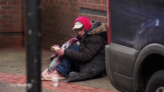 Eine krank aussehende Frau sitzt im Regen auf der Straße. Vor ihr steht ein Pappbecher. © Screenshot 