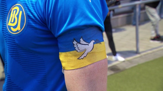 Nahaufnahme auf einen Mann, der ein blaues Trikot trägt. Als Kapitänsbinde trägt er eine Binde in den ukrainischen Nationalfarben auf der eine weiße Taube drauf ist. © Screenshot 