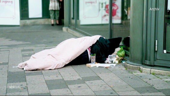 Ein obdachloser Mensch liegt auf der Straße an einer Häuserwand. © Screenshot 