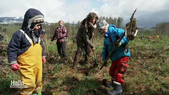 Kinder mit einer Schaufel vor einer Wieder mit Menschen die Bäume pflanzen. © Screenshot 
