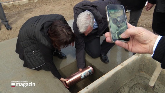 Zwei Personen legen eine Kupferkapsel in ein Loch in der Erde. Eine andere Person dokumentiert mit dem Smartphone. © Screenshot 