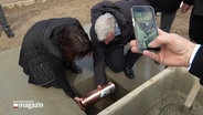 Zwei Personen legen eine Kupferkapsel in ein Loch in der Erde. Eine andere Person dokumentiert mit dem Smartphone. © Screenshot 