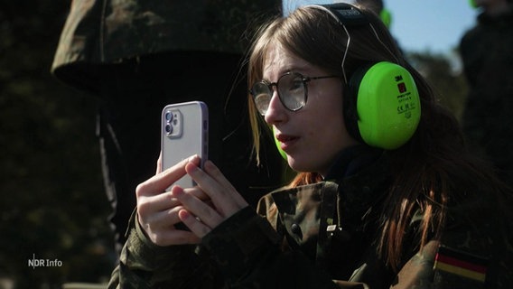 Eine Schülerin in Tarnkleidung, mit Gehörschutz und Smartphone. © Screenshot 