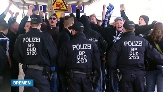 Pöbelnde Fußballfans auf einem Gleisabschnitt, mit dem Rücken zum Betrachter davor Polizisten in Schutzkleidung und mit Ausrüstung. (Archivbild) © Screenshot 