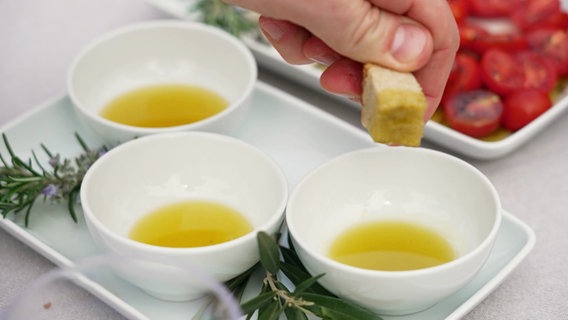 Auf einem Tisch stehen drei weiße Schälchen mit Olivenöl und frische Tomaten. Jemand tunkt ein Stück Brot in das Öl. © Screenshot 