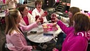 Fünf Frauen in pink-rosa Kleidung sitzen an einem Tisch und stoßen zu Popcorntüten mit ihren Getränken an. © Screenshot 