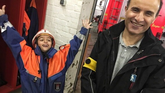 Ahmad steht lächelnd neben seinem Sohn, der in Feuerwehr-Montur begeistert die Hände in die Luft streckt.  