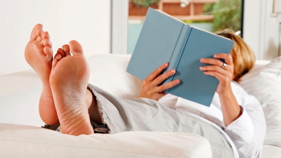 Eine Frau liegt auf dem Sofa, hat ihre Füße hochgelegt und liest ein Buch. © picture alliance / Klaus Ohlenschläger Foto: Klaus Ohlenschläger