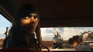 Eine Frau sitzt im dunklen Auto, nur ihre Augen sind hell erleuchtet - im Hintergrund ein Tankwagen - Szene aus "Furiosa" © 2023 Warner Bros. Entertainment Inc. All Rights Reserved. Foto: Jasin Boland