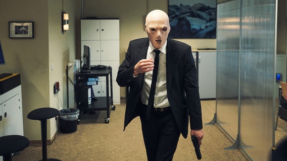 Ein Mann im Anzug läuft mit einer Pistole in der Hand und Maske über dem Gesicht durch einen Raum in einer Bank. © ARD Degeto Foto: Marco Fischer