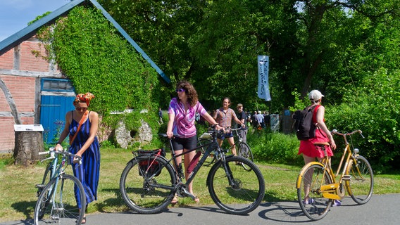 Drei sommerlich gekleidete Frauen mittleren Alters schieben ihre Fahrräder entlang einer Straße. Hinter ihnen sind weitere Menschen und ein Bauernhaus oder eine Scheine in rotem Backstein, mit Efeu bewachsen. © www.kulturelle-landpartie.de 