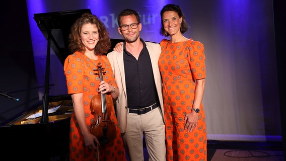 Ein Mann steht zwischen zwei Frauen, die in orange gekleidet sind. © Claudius Hinzmann / NDR Foto: Claudius Hinzmann / NDR