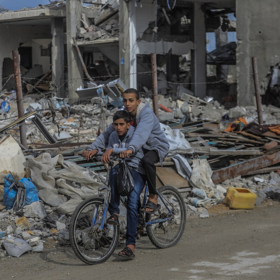 Ein Palästinenser mit einem Kind fährt auf einem Fahrrad durch eine Straße mit zerstörten Häusern © picture alliance/dpa Foto: Mohammed Talatene