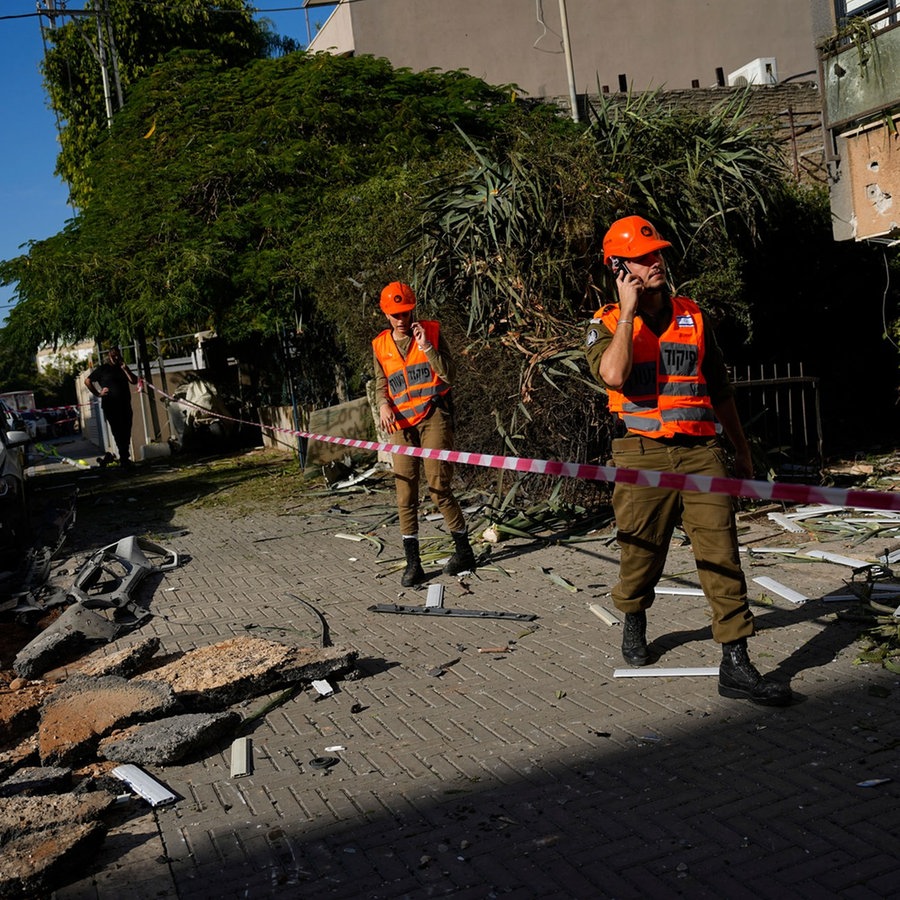 Israelische Sicherheitskräfte stehen an einer Stelle auf einer beschädigten Straße und vor mehreren kaputten Autos, die von einer aus dem Gazastreifen abgefeuerten Rakete getroffen wurde. © picture alliance/dpa/AP Foto: Ariel Schalit