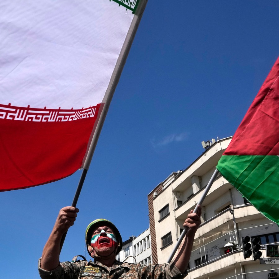 Ein Mann, dessen Gesicht in den Farben der iranischen Fahne bemalt ist, schwenkt links die iranische und rechts die palästinensische Fahne. © AP/dpa Foto: Vahid Salemi