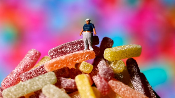 Ein übergewichtiger Mann steht auf einem Berg Süßigkeiten. (Symbolbild Zuckerkonsum) © picture alliance / Panama Pictures | Christoph Hardt 