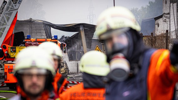 Zwei Feuerwehrleute unterhalten sich und im Hintergrund sind verbrannte Überreste einer Halle zu sehen. © dpa-Bildfunk Foto: Moritz Frankenberg/dpa
