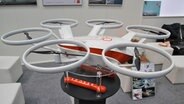 At CeBIT 2016, you can see a white drone with a red cross in a hall of the Hannover exhibition center. On red feet is "Rescue". © NDR Photo: Jürgen Jenauer