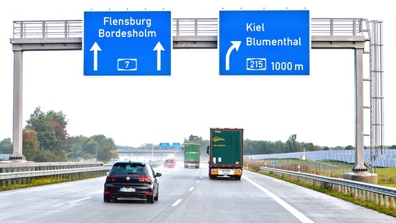Blick auf ein großes Autobahnschild mit der Abzweigung rechts nach Kiel/Blumenthal in 1000m und geradeaus in Richtung Flensburg/Bordesholm. Unter dem Schild fahren links ein Auto und rechts ein LKW. © picture alliance/dpa Foto: Frank Molter