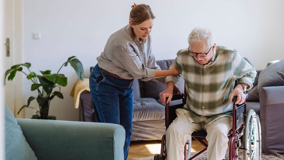 Eine junge Frau hilft einem älteren Herr aus dem Rollstuhl. © IMAGO / Westend61 