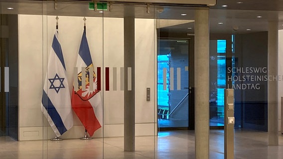 Die Israelische und Schleswig-Holsteinische Flagge stehen nebeneinander hinter einer Glaswand im Landtag. © NDR Foto: Anna Grusnick