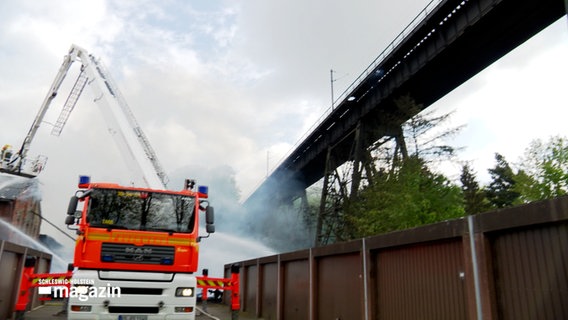 Eine Drehleiter der Feuerwehr ist auf ihren Stützen aufgebockt, Feuerwehrleute löschen ein Feuer neben der Eisenbahnhochbrücke in Rendsburg © NDR Foto: NDR Screenshot