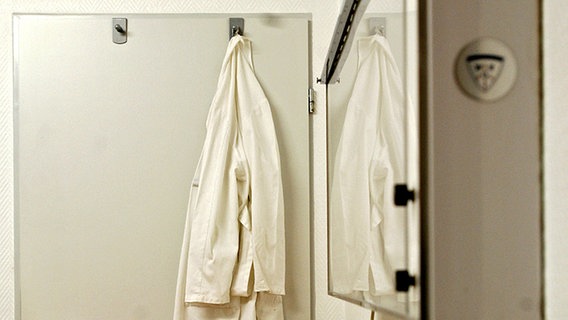 Ein weißer Arztkittel hängt in einer Arztpraxis an einem Haken an der Tür. © dpa - Report Foto: Frank May
