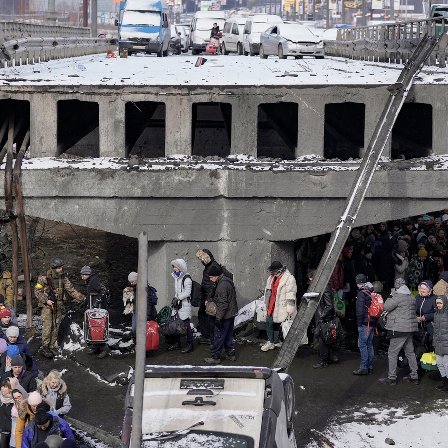 Ukrainische Männer und Frauen stehen auf der Flucht aus Irpin vor einer zerstörten Brücke, auf der verlassene, eingeschneite Autos stehen. © AP/dpa Foto: Vadim Ghirda