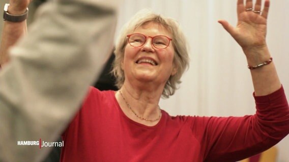 Eine ältere Frau im roten Pullover lächelt während sie beim Tanzen ihre Arme in die Höhe streckt. © Screenshot 