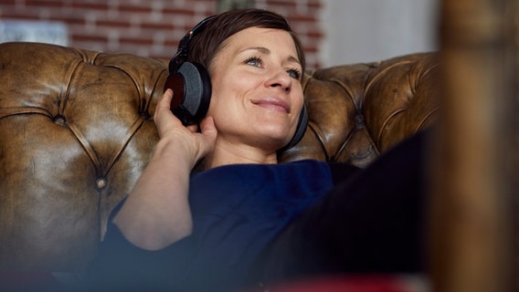 Frau liegt lächelnd mit Kopfhörern auf dem Sofa und hört Musik. © photocase.de Foto: Westend61