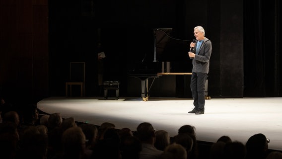 Auf einer Bühne steht ein weißhaariger Mann in schwarzer, legerer Kleidung. Es ist der ehemalige Ballettchef des Hamburg Ballett John Neumeier. © Hamburg Ballett / Kiran West Foto: Kiran West