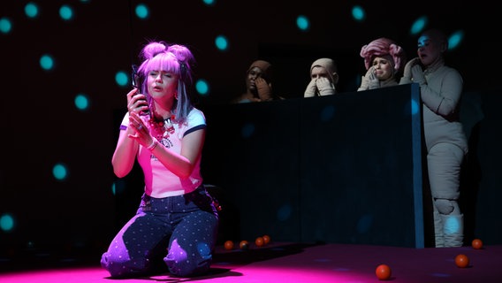 Ein junge Frau mit rosa Comic - Manga Zöpfen kniet auf einem schwarzen Boden und schaut in ihre handy. © Staatsoper Hamburg Foto: Jörg Landsberg