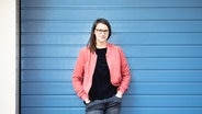 Eine junge Frau mit langem breunem Haar und Brille in einer rosa Jacke vor einer blauen Wand. © Sebauer.Filmer - DuMont Verlag Foto: Sebauer.Filmer