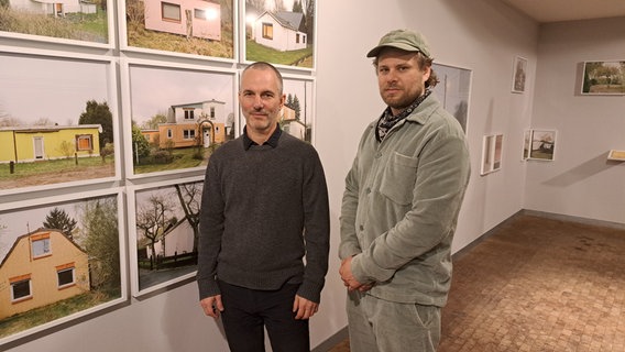 Vor Fotos von Behlfsheimen, sogenannten "Ley- Buden" stehen zwei Männer. Es sind die Fotografen Enver Hirsch und Philipp Meuser. © NDR Foto: Petra Volquardsen