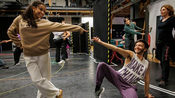 In einem Probenraum reicht eine Frau lächelnd einem jungen Mann die Hand, der neben ihr kniet. © StageEntertainment Foto: StageEntertainment