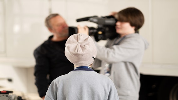 Ein Kind steht vor einem anderen Kind und einem Erwachsenen, die zusammen eine Kamera bedienen. © NDR Foto: Jann Wilken