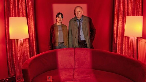 Almila Bagriacik (r.) und Axel Milberg stehen bei einer Szene im Tatort "Borowski und das hungrige Herz" hinter einem roten Sofa © NDR/Thorsten Jander Foto: Thorsten Jander