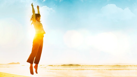 Eine Frau springt vor der untergehenden Sonne am Strand in die Luft.  