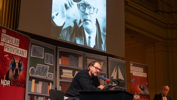 Charly Hübner sitzt auf einer Bühne und liest aus einem Buch. © Silke Winkler Foto: Silke Winkler