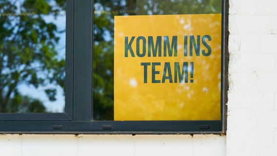 Ein gelbes Schild mit der Aufschrift "komm ins Team" steht in einem Fenster © Colourbox Foto: Heiko Küverling